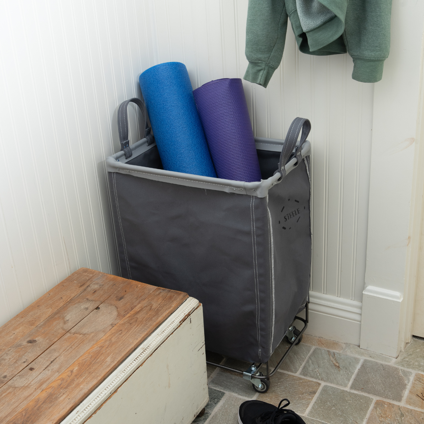 Gray cart with blue and purple yoga mats inside, next to a wooden bench and green jacket on a white wall.