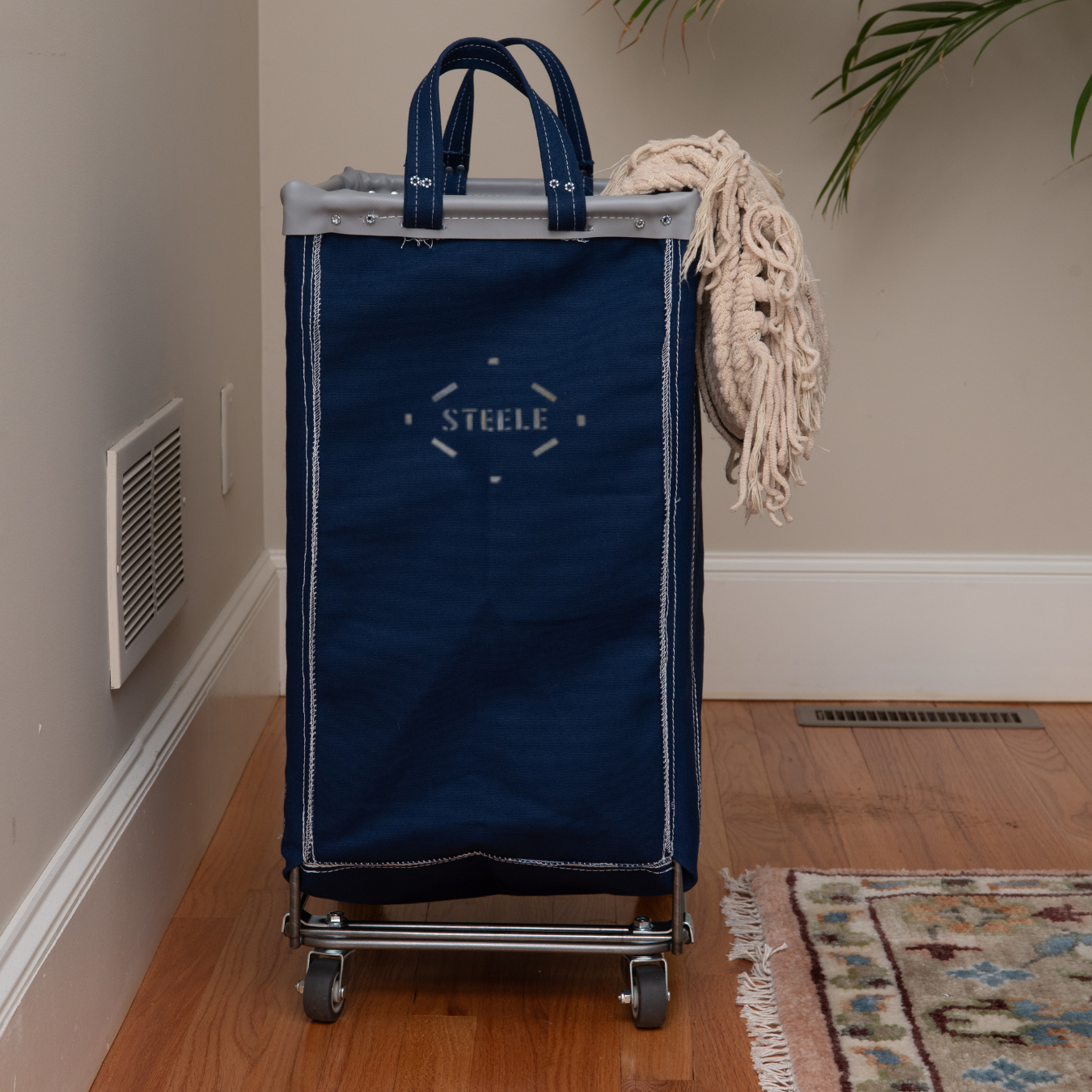 Blue STEELE rolling laundry bag on wheels in a room with wooden floor and plant.