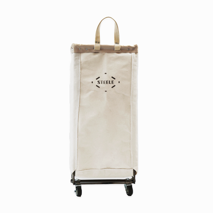 A tall canvas laundry hamper on wheels by STEELE, in a natural off-white canvas color with a steel base and handles, shown against a plain white background.