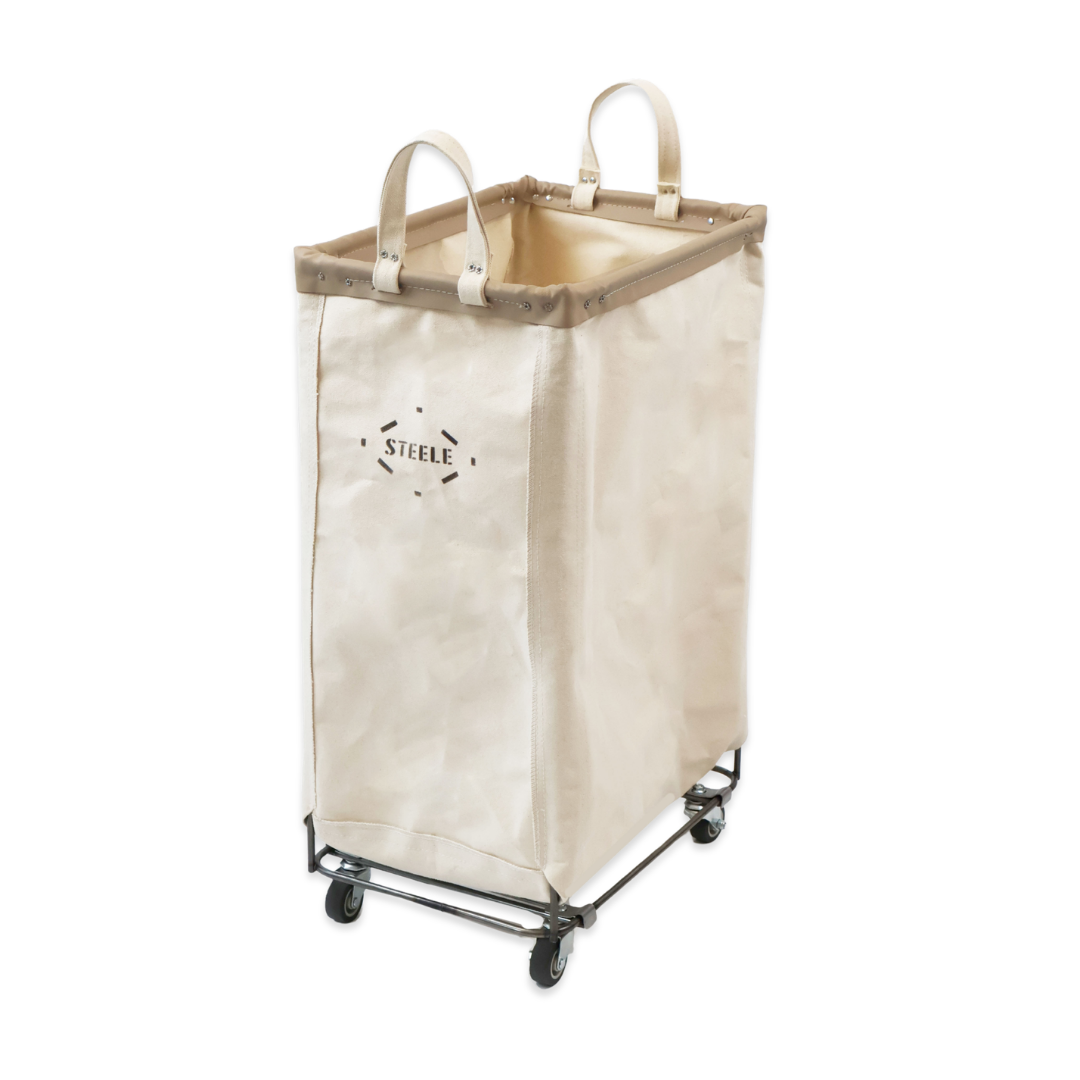 A tall canvas laundry hamper in a warm beige color with dual top handles, a steel wheeled frame, and a black 'STEELE' logo printed on the front, set against a plain white background.