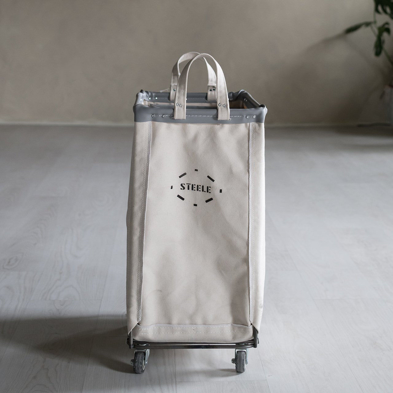 Beige rolling hamper with 'STEELE' branding on a gray floor.