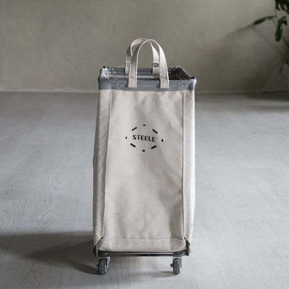 Beige rolling hamper with 'STEELE' branding on a gray floor.