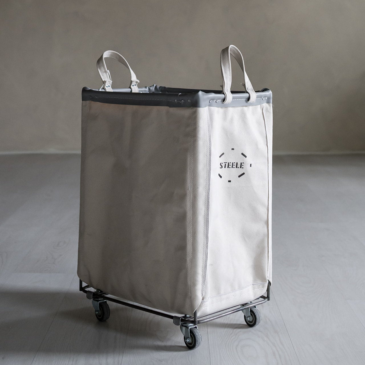 natural canvas laundry cart with wheels on a gray floor against a plain wall