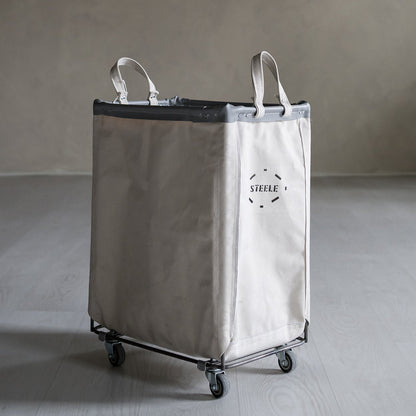 natural canvas laundry cart with wheels on a gray floor against a plain wall