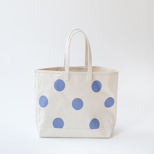 A cream canvas tote bag with large blue polka dots and matching cream shoulder straps, photographed against a seamless white studio background.