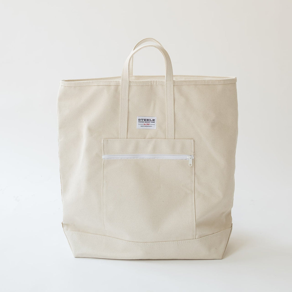 A light beige canvas tote bag with a front zippered pocket and a small white brand label, photographed against a plain off-white background.