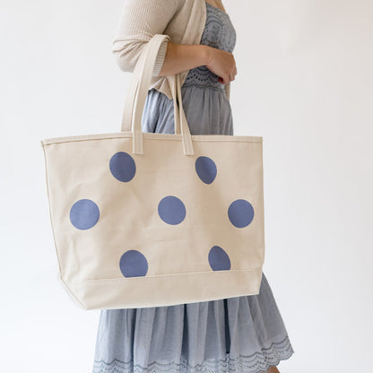 A person wearing a light gray dress holds a large cream canvas tote bag with blue polka dots against a plain white background.