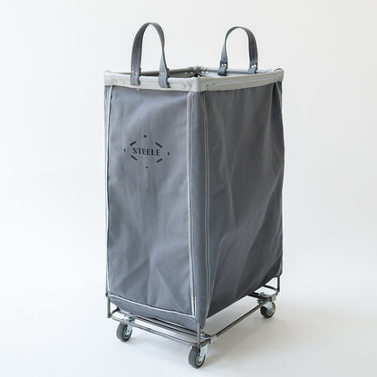 A gray fabric laundry hamper with dual top handles, a metal rolling frame, and caster wheels, shown on a clean white studio background. The brand name 'STEELE' is printed on the front.