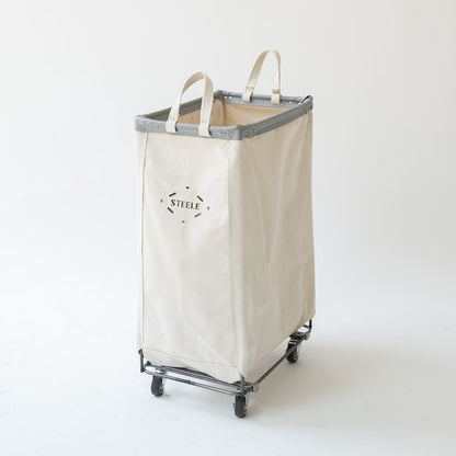 A tall canvas laundry hamper in a warm off-white color with a gray metal frame and black caster wheels. The front features a black printed logo that reads 'STEEL'.