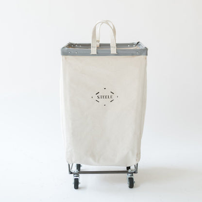 A tall canvas laundry hamper with a gray metal frame and wheels, featuring a top handle and a black 'STEELE' logo printed on the front, set against a plain white background.