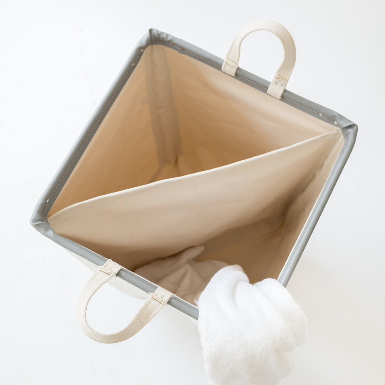 A square fabric storage bin with a gray exterior and a beige interior, shown open on a white background with a white towel hanging from one handle.