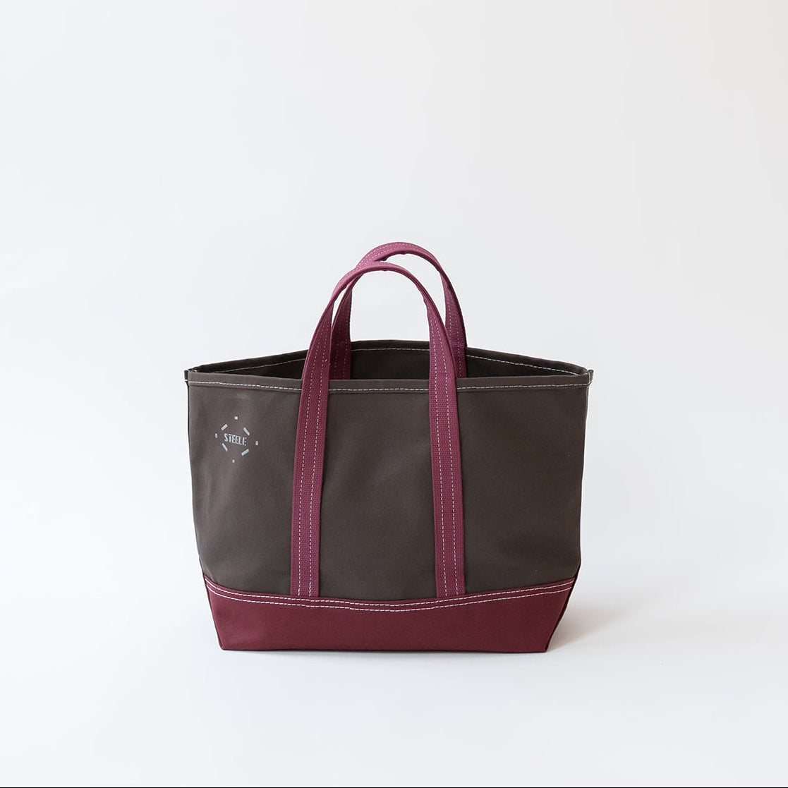 A dark brown canvas tote bag with a deep burgundy base and matching burgundy handles, shown against a plain white studio background.