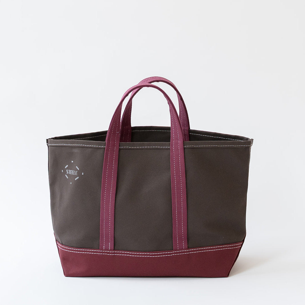 A canvas tote bag in dark brown with magenta-pink handles and bottom trim, featuring a small white embroidered logo on the left side, set against a plain white background.