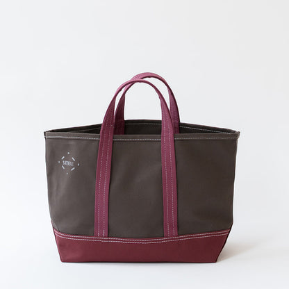 A canvas tote bag in dark brown with magenta-pink handles and bottom trim, featuring a small white embroidered logo on the left side, set against a plain white background.