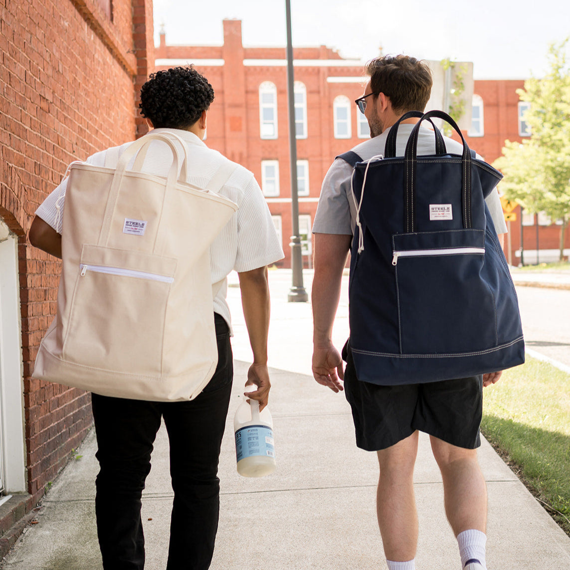 Two people walking on a sidewalk with backpacks, one beige and one navy, in front of a brick building.