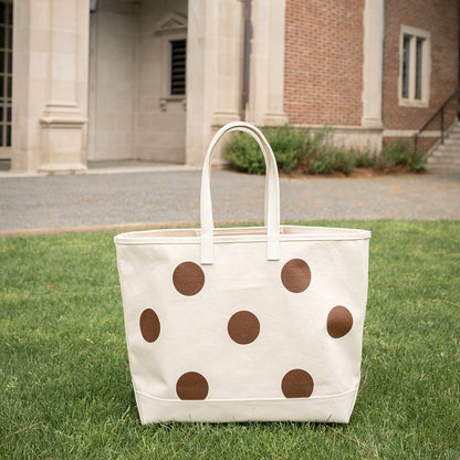 White tote bag with brown polka dots on grass in front of a building