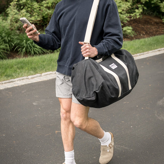 A man walking outdoors carrying a large black duffel bag with cream-colored straps and trim. He is wearing a navy sweatshirt, light gray shorts, white socks, and beige slip-on shoes.