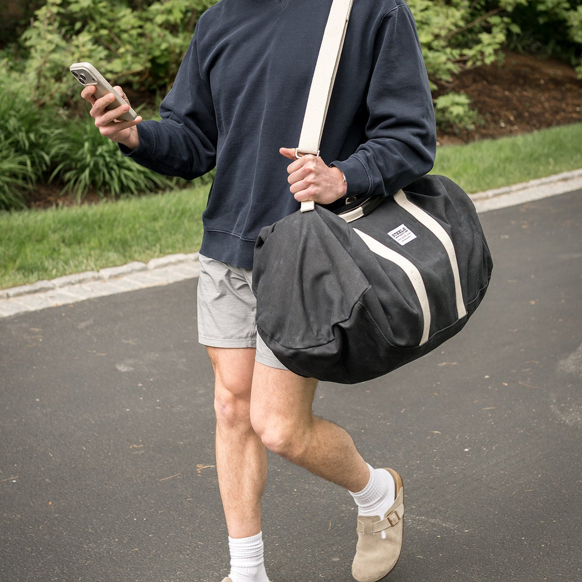 A man walking outdoors carrying a large black duffel bag with cream-colored straps and trim. He is wearing a navy sweatshirt, light gray shorts, white socks, and beige slip-on shoes.