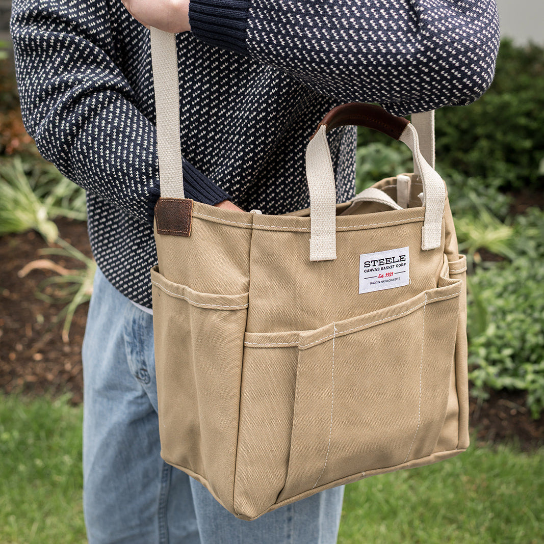 A person outdoors wearing a dark patterned sweater and jeans, carrying a beige canvas utility tote bag with cream straps and brown leather-like handles. The bag has front pockets and a visible STERLO label.
