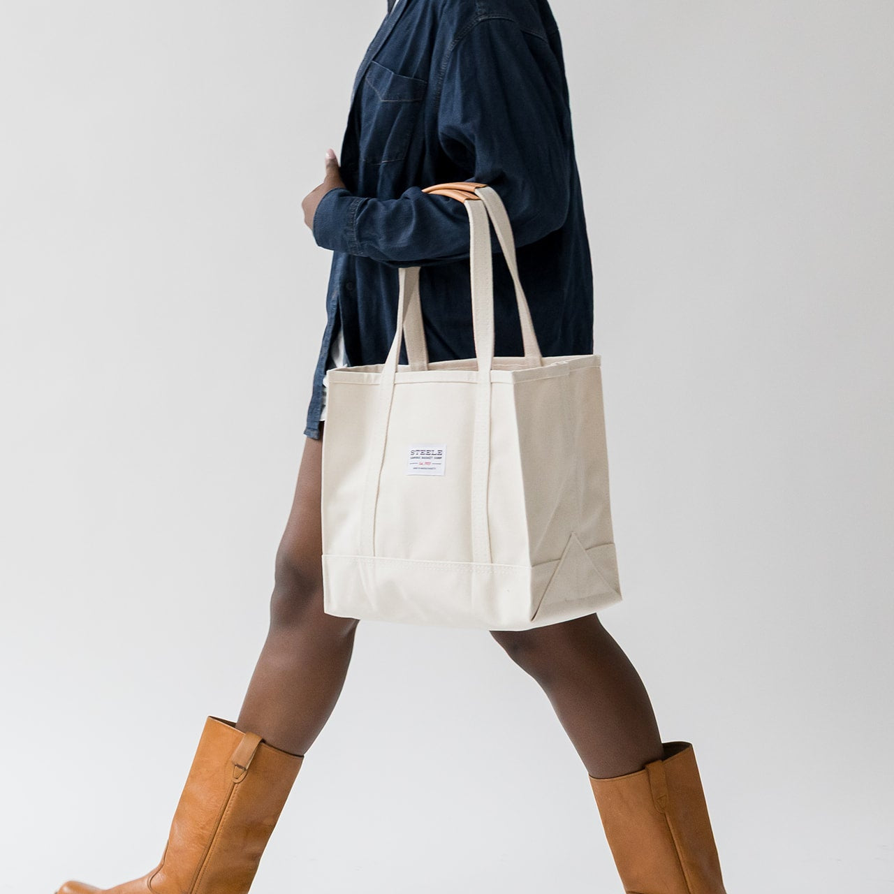 Person holding a white tote bag with a visible brand logo, wearing brown boots on a plain background