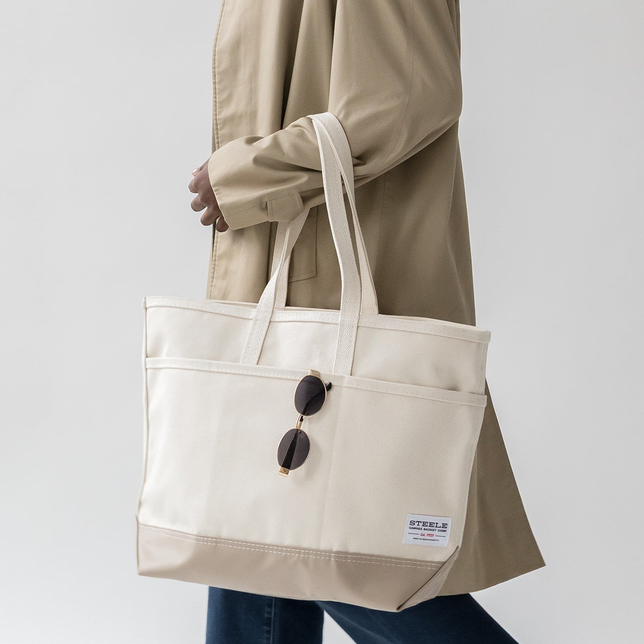 Person holding a beige tote bag with sunglasses on a plain background