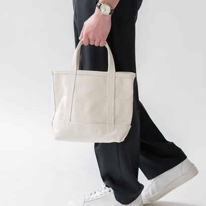 Person holding a beige tote bag wearing black pants and white sneakers on a light gray background