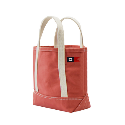A nantucket red canvas tote bag with cream-colored fabric handles and a small red, black, and white logo patch on the front, set against a plain white background.