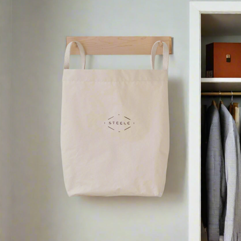 Beige tote bag hanging on a wooden hook against a light gray wall with clothes in the background.