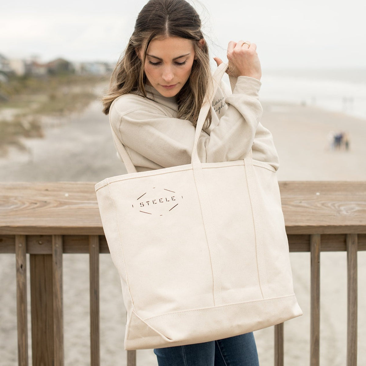 A woman at the beach holding a beige canvas tote bag with the brand 'ARTIE' visible on the side.