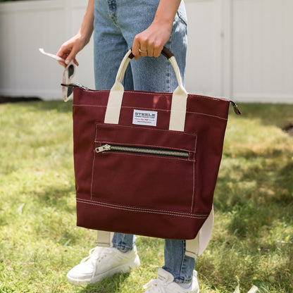 Person holding a maroon tote bag with a visible brand label in an outdoor setting.
