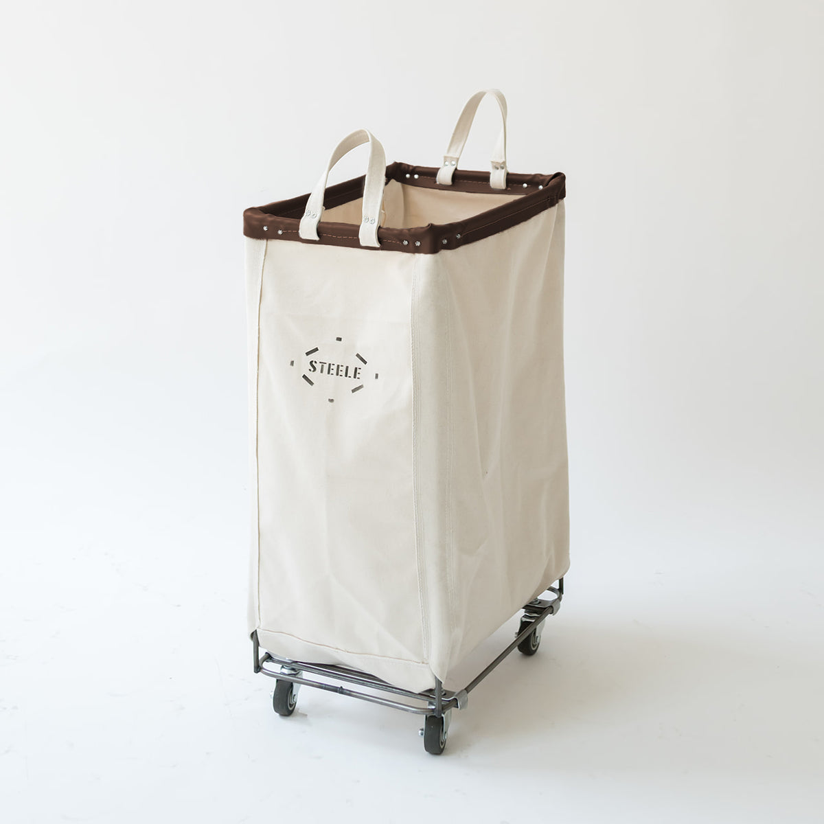 A canvas laundry hamper with a dark brown leather trim, and a metal rolling frame with wheels. The brand name 'STEELE' is printed on the front.