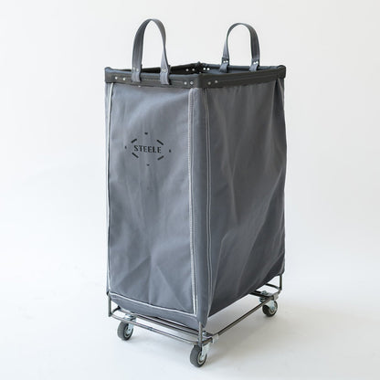 A gray canvas laundry hamper with dual top handles, a metal frame, and four caster wheels. The front features a black logo that reads 'STEELE'.