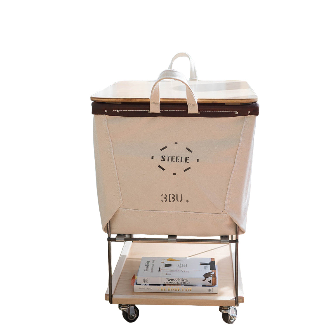 A natural canvas laundry hamper with a steel rolling cart base, featuring a lid, top handles, and a lower shelf holding a magazine, on a plain white background.