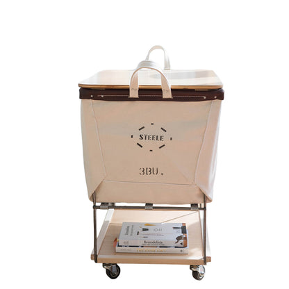 A natural canvas laundry hamper with a steel rolling cart base, featuring a lid, top handles, and a lower shelf holding a magazine, on a plain white background.
