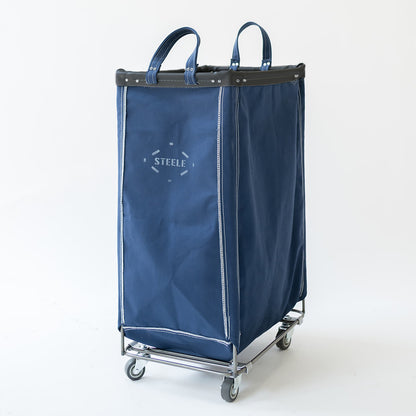 A navy canvas laundry hamper with dual top handles, a black leather trim, and a silver metal wheeled base. The front features a white printed logo that reads 'STEELE'.