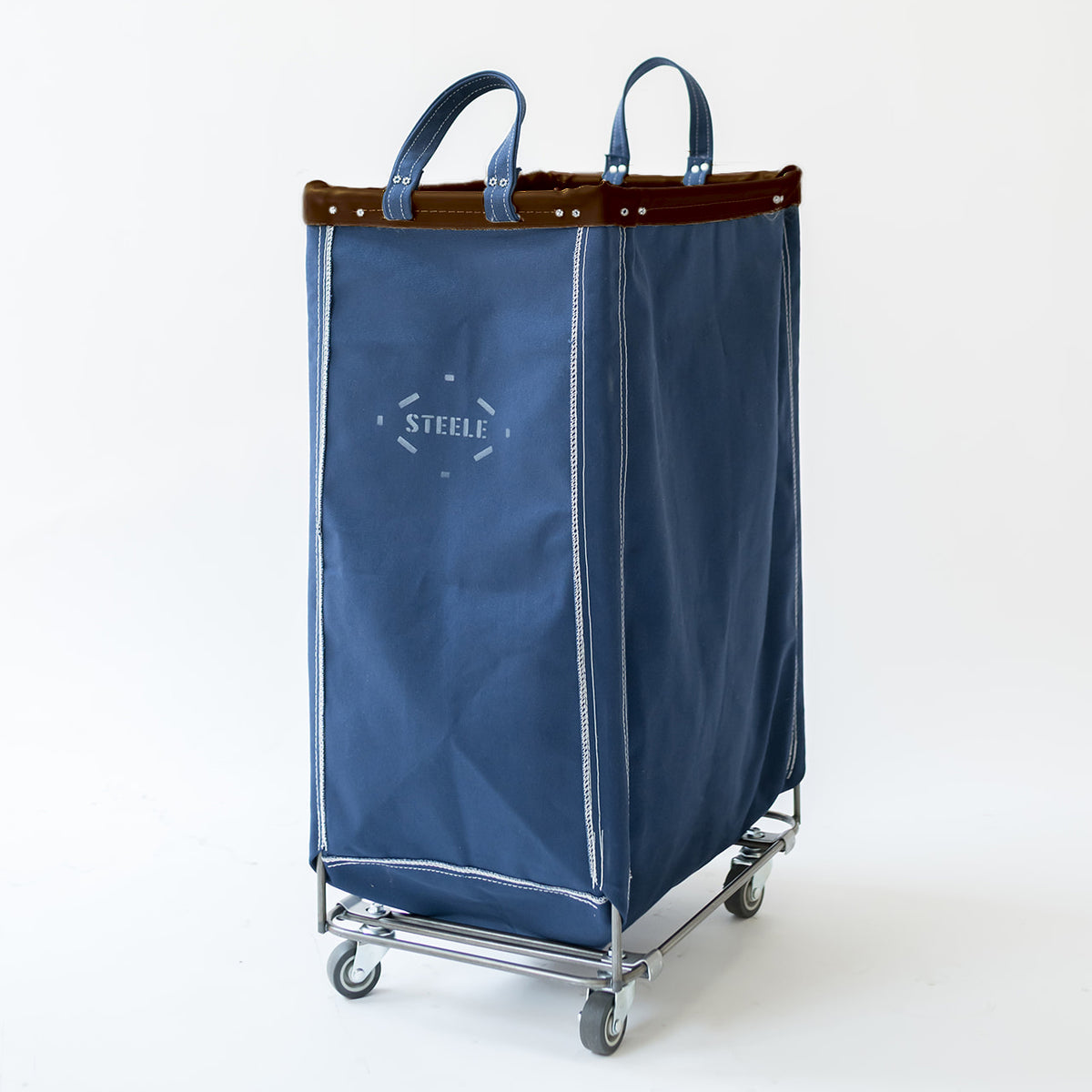 A navy blue canvas laundry hamper with a brown leather trim, blue handles, and a silver metal frame with caster wheels. The front features a white embroidered logo that reads 'STEELE'.