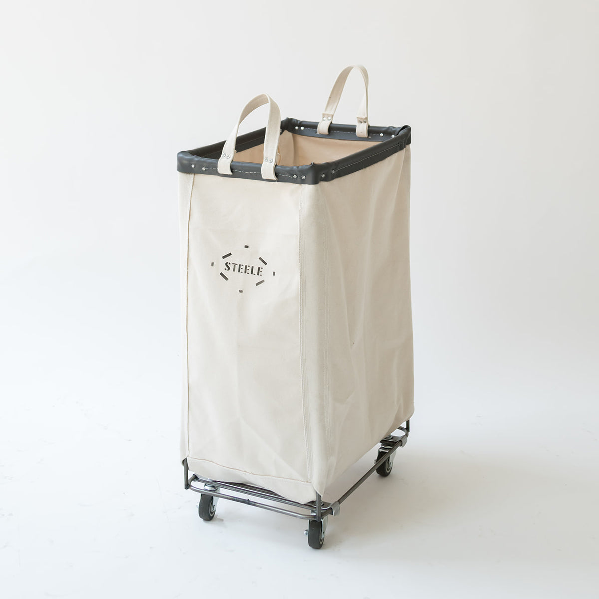 A natural canvas laundry hamper with black leather trim and handles, mounted on a metal rolling frame with wheels. The front features the brand name 'STEELE' printed in black.