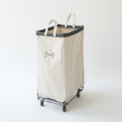 A natural canvas laundry hamper with black leather trim and handles, mounted on a metal rolling frame with wheels. The front features the brand name 'STEELE' printed in black.
