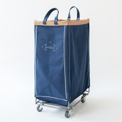 A navy canvas laundry hamper with dual top handles, a vegetable tan leather top rim, and a silver metal wheeled base. The front features a white printed logo that reads 'STEELE'.