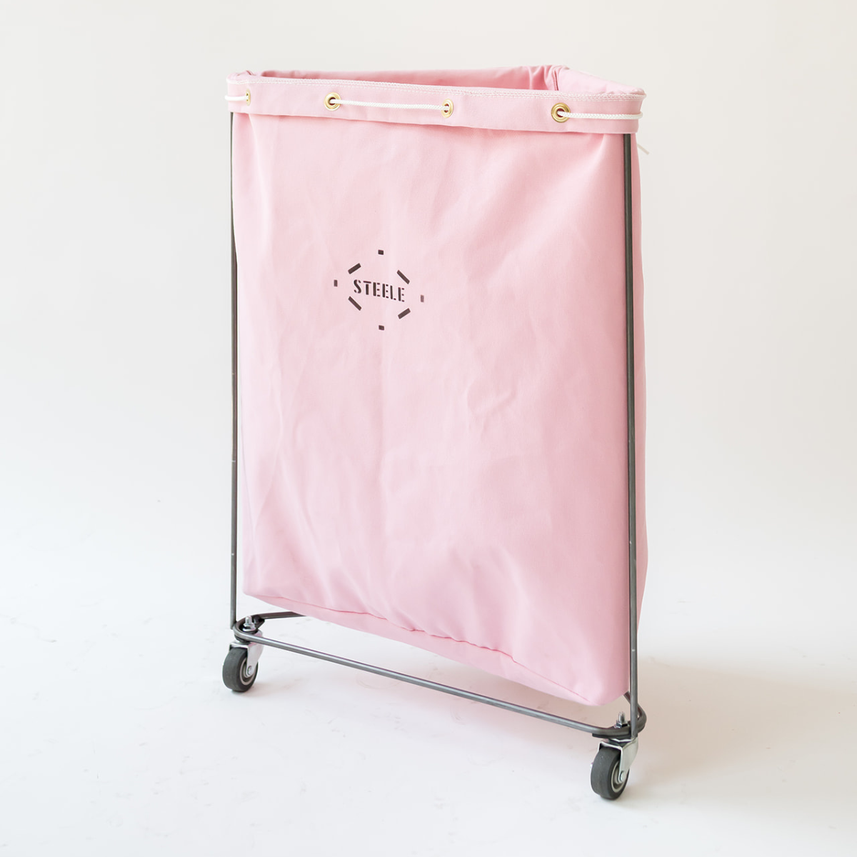 A pink canvas laundry hamper with a metal frame and caster wheels, featuring the brand name 'STEELE' printed in black on the front.