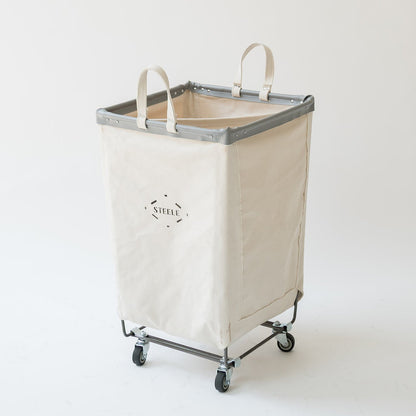 A tall canvas laundry hamper with a cream-colored body, gray metal frame, and four caster wheels, featuring dual top handles and a 'STELLA' logo on the front.