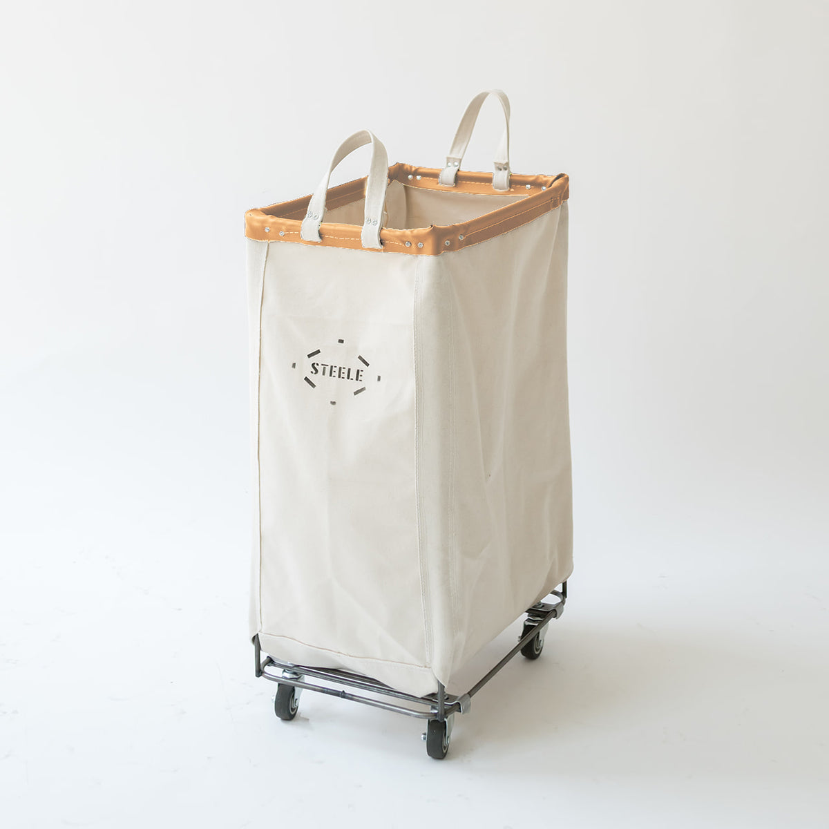 A canvas laundry hamper with a light vegetable tan leather trim at the top, dual fabric handles, and a metal wheeled base. The front features a black printed logo that reads 'STEELE'.