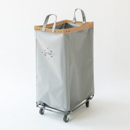 A light gray vinyl laundry hamper with a vegetable tan leather trim, metal handles, and a wheeled base, shown on a plain white studio background.