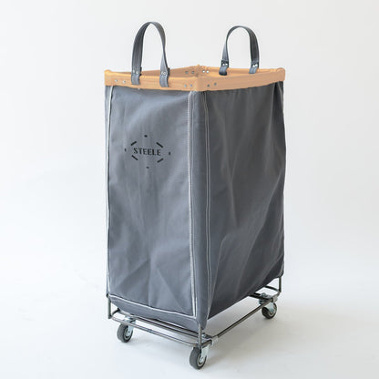 A gray canvas laundry hamper with a vegetable tan leather trim, dual gray canvas handles, and a steel rolling frame with four wheels. The word 'STEELE' is printed on the front.