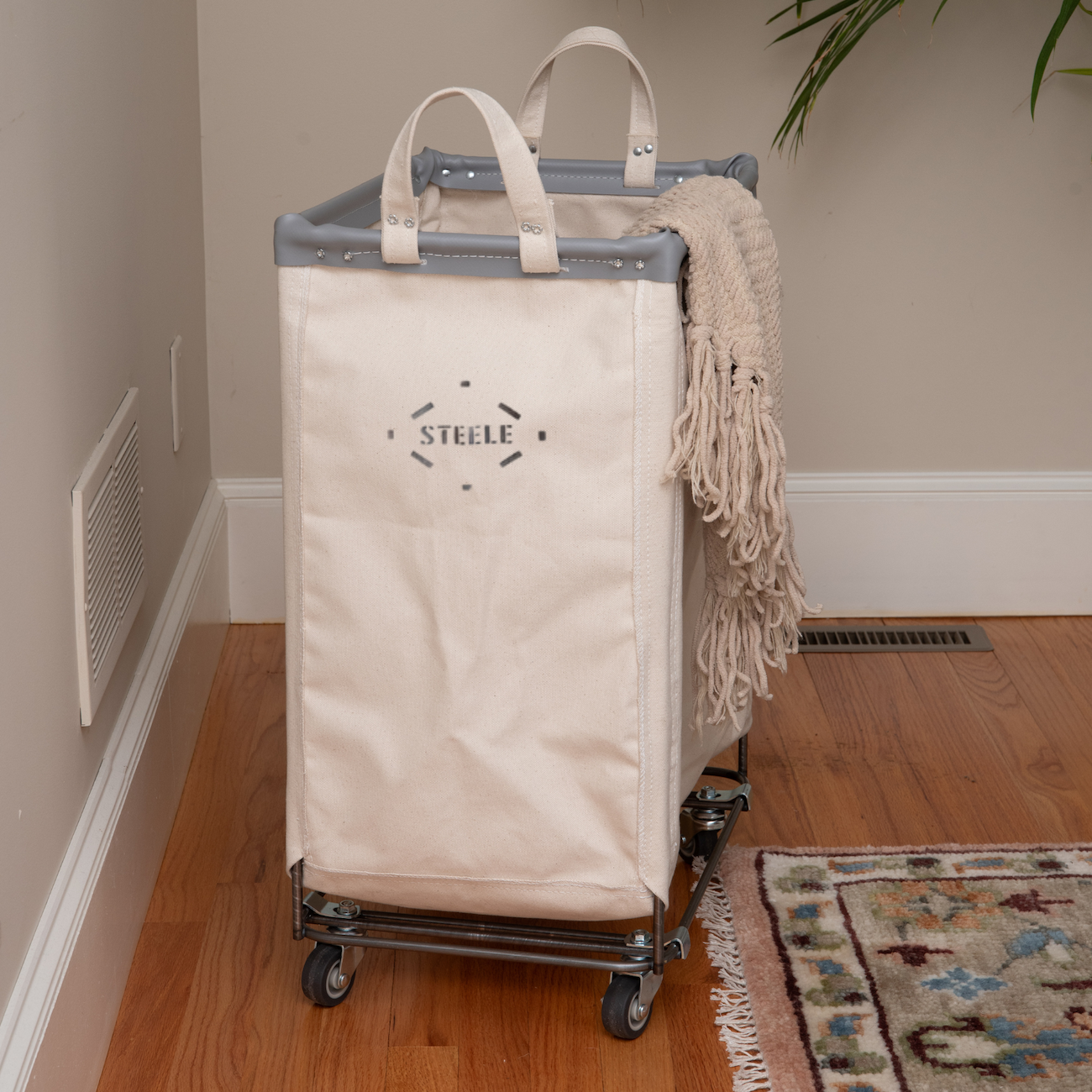 Natural laundry cart with 'Steele' branding on wheels in a room.