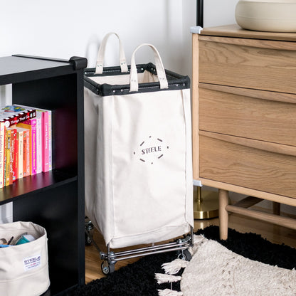 White laundry cart with 'STEELE' branding next to a wooden dresser and bookshelf.
