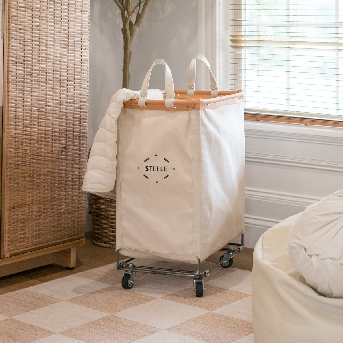 Beige laundry basket with wooden handle and 'Steele' branding in a room setting.