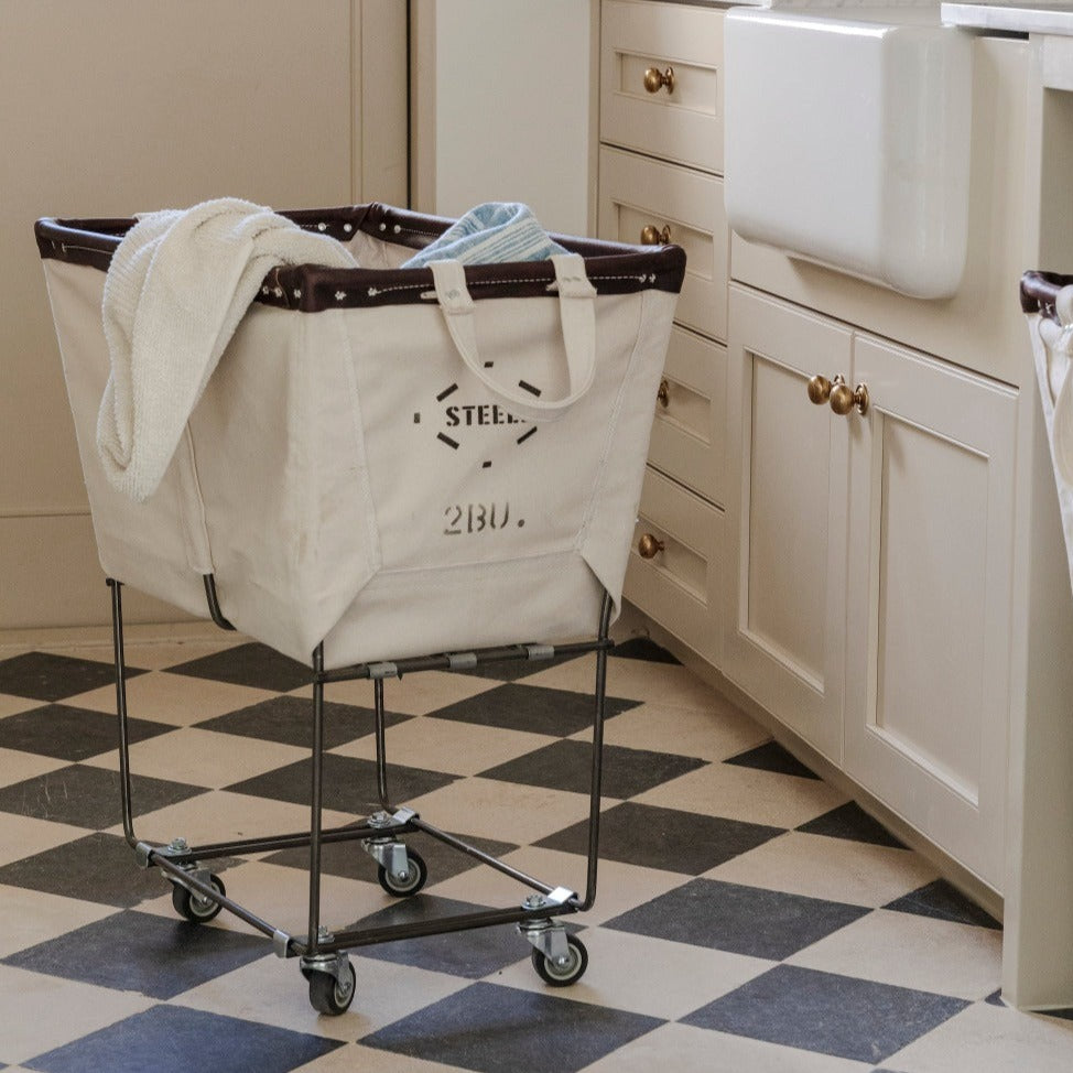 A canvas canvas laundry sorter on wheels, with 'STEELE 2BU' printed on the side, placed in a kitchen setting.