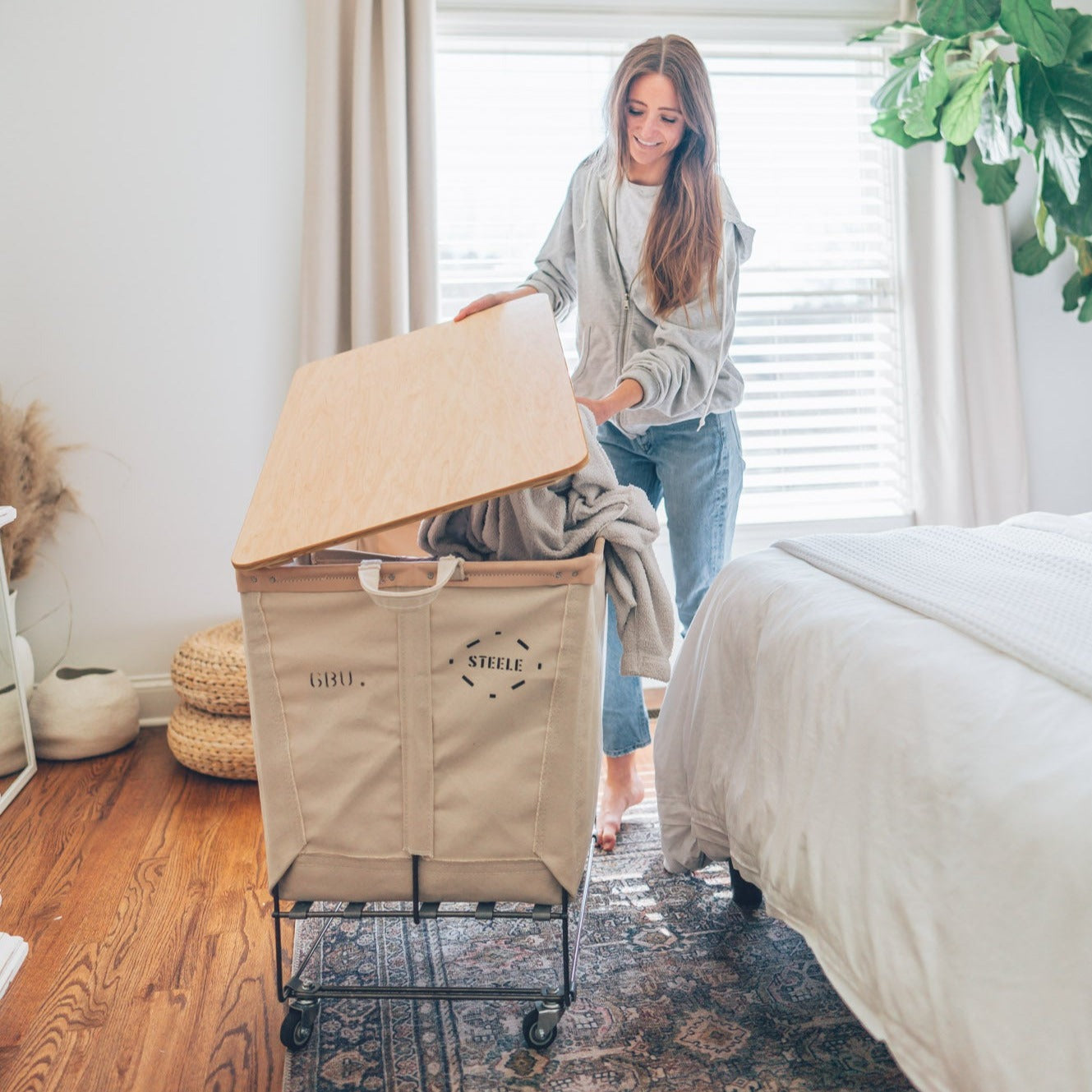 A woman is interacting with a natural canvas laundry sorter truck with black canvas dividers in a room with a bed and plants. The truck has a metal frame and wheels.
